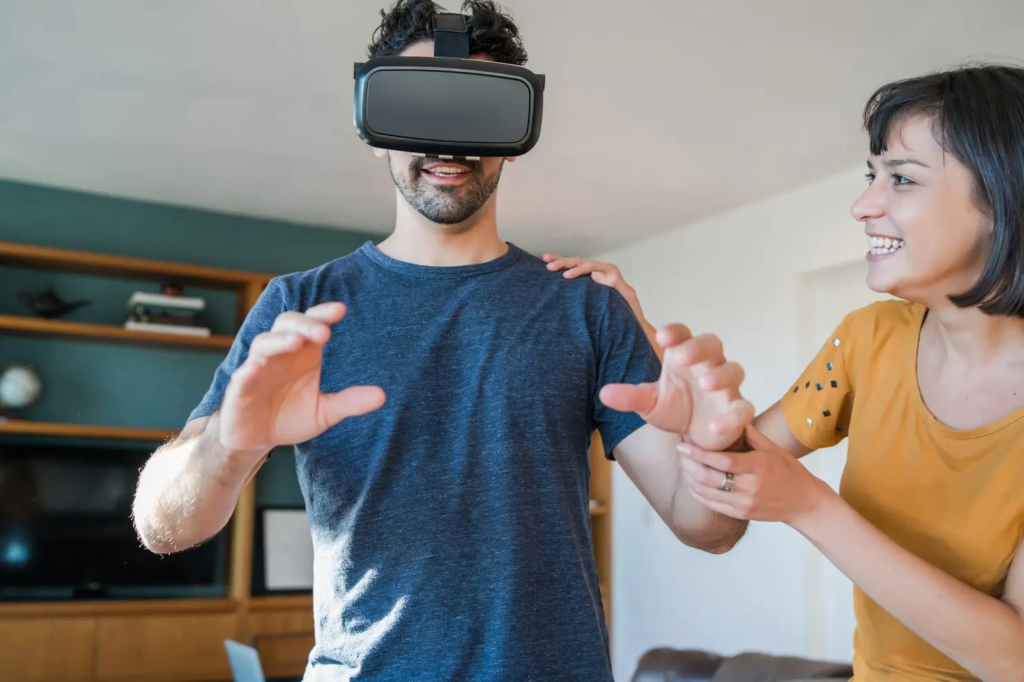 Man wearing a virtual reality headset reaches forward with both hands while a woman stands beside him and smiles