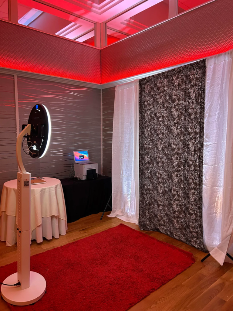 Photo booth setup at a Mitzvah event with a ring light camera, printer, patterned backdrop, white curtains, and red carpet