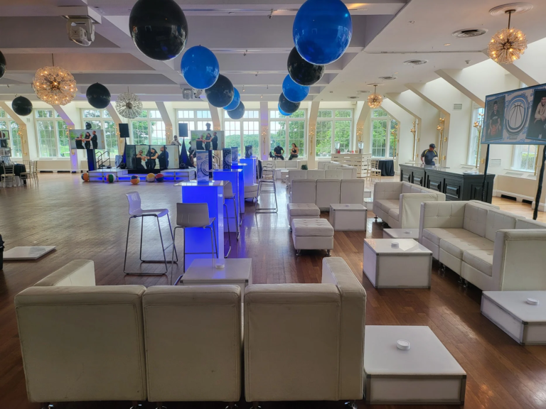 Spacious event venue featuring white lounge sofas, ottomans, coffee tables, high-top seating, and blue and black balloons arranged around a dance floor