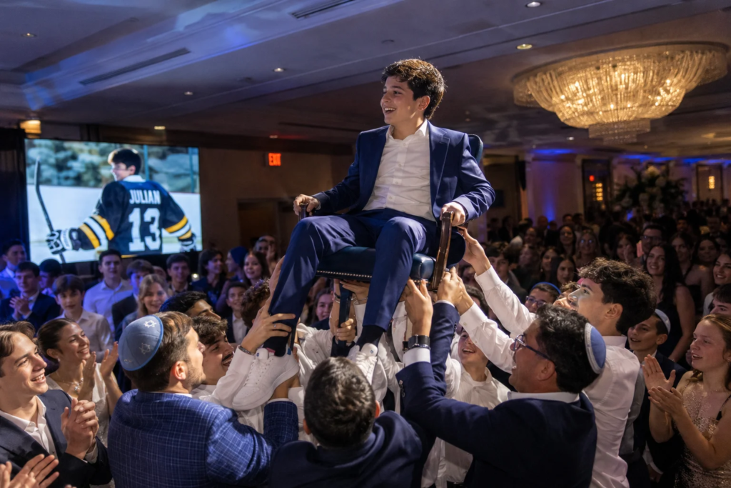 Teen boy in a blue suit is lifted on a chair by guests during a lively Mitzvah celebration, with a crowd dancing and cheering around him