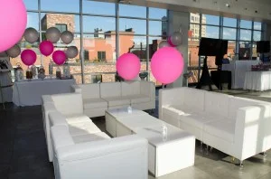White sofas, armchairs, and square coffee tables arranged in a lounge seating area inside a bright event venue with colorful balloons
